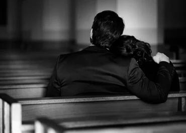 A man and a woman sitting inside the church they both wear a black suit, the womans head is leaning on a mans shoulder, they are both in grief.